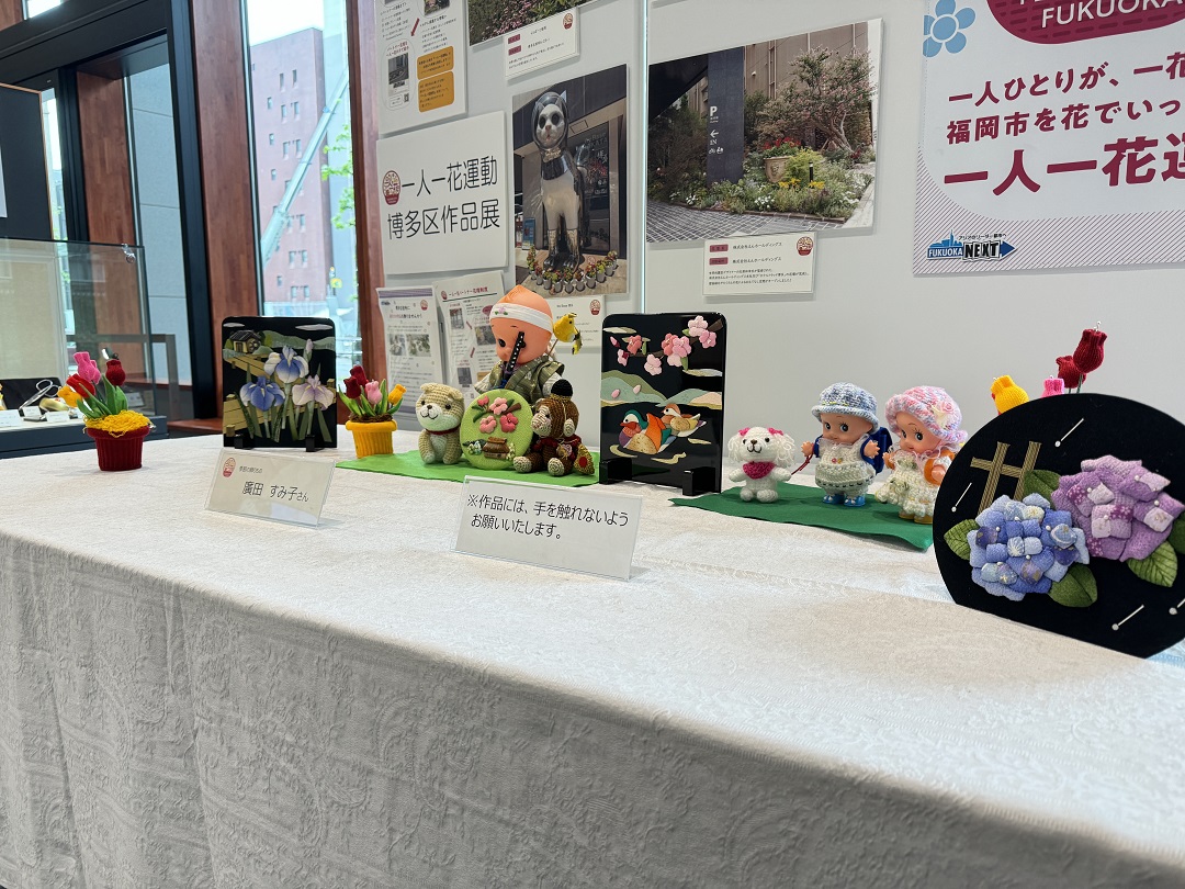 作品展全体の様子の写真。菖蒲の花、桜の花と里山、湖面に散る桜の花と鳥の親子、紫陽花の花の四点の切り込み作品とチューリップなどの編み物の作品が並んでいる。