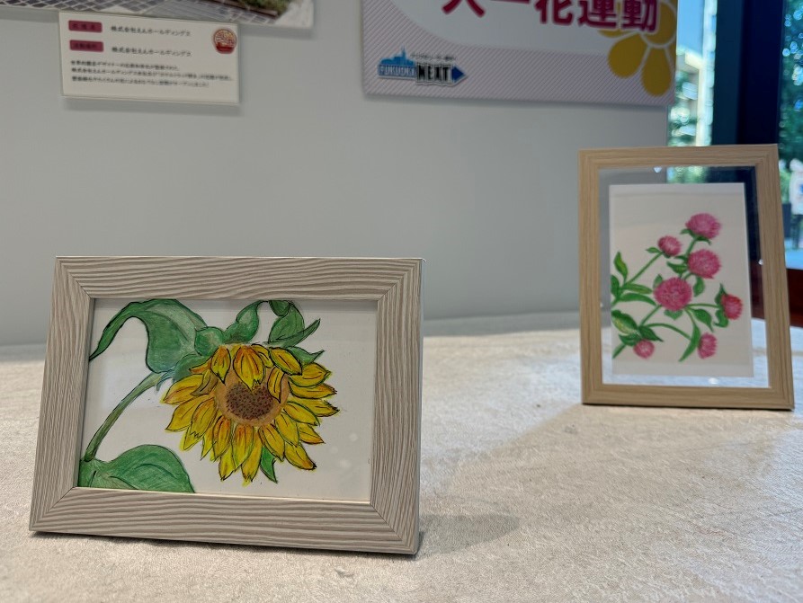 一人一花運動博多区作品展の写真。木製のフレームに入ったひまわりのイラストが飾られている。