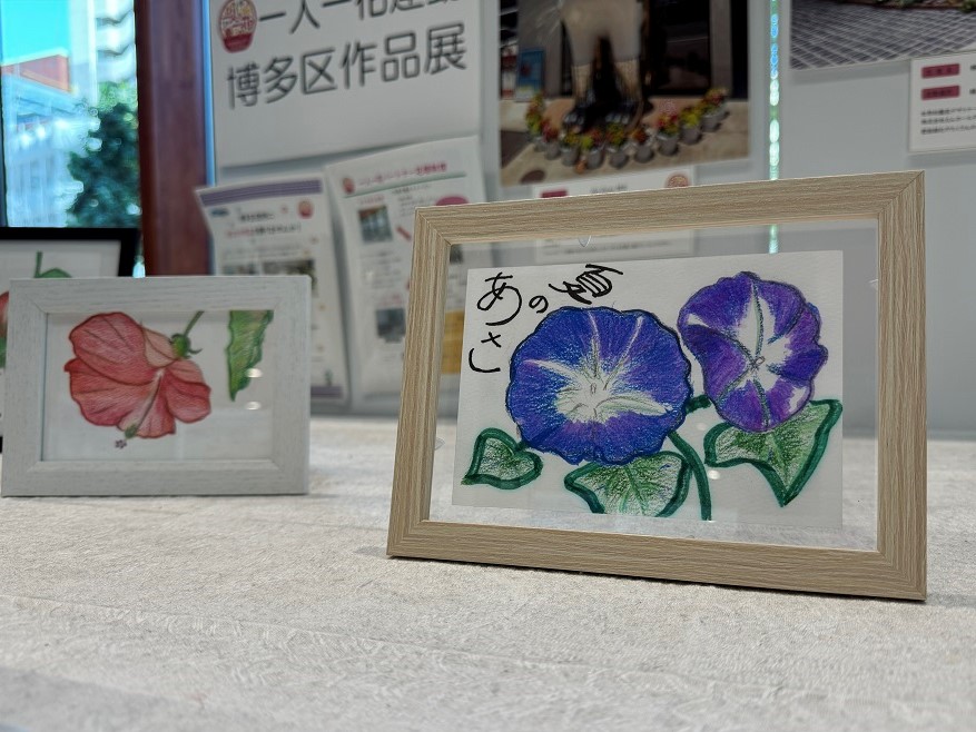 一人一花作品展の写真。木製のフレームに入った青紫色の朝顔の花のイラストが飾られている。