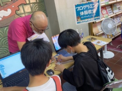 子ども二人がスタッフに教えられながらプログラミングを体験している写真