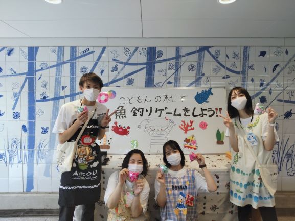 魚釣りゲームと書かれた白板の前で、学生４人が写っている写真