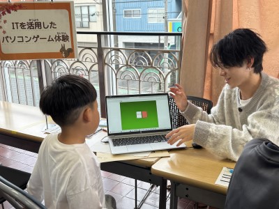 学生が、小学生に学生自身がプログラミングしたゲームを教えている画像