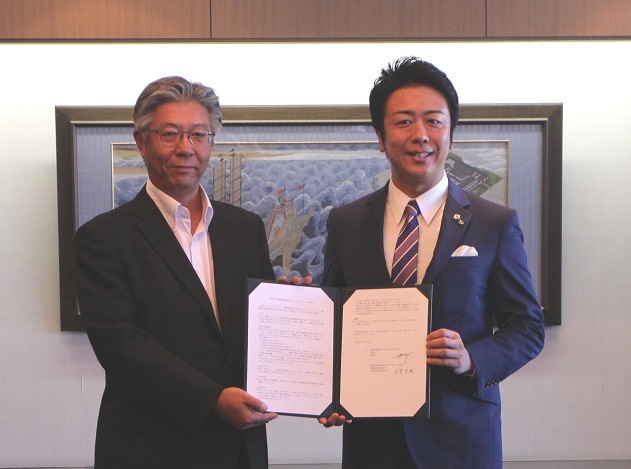 福岡国際空港株式会社永竿代表取締役社長執行役員と高島市長がパートナーシップ協定書を持ち，ツーショット撮影
