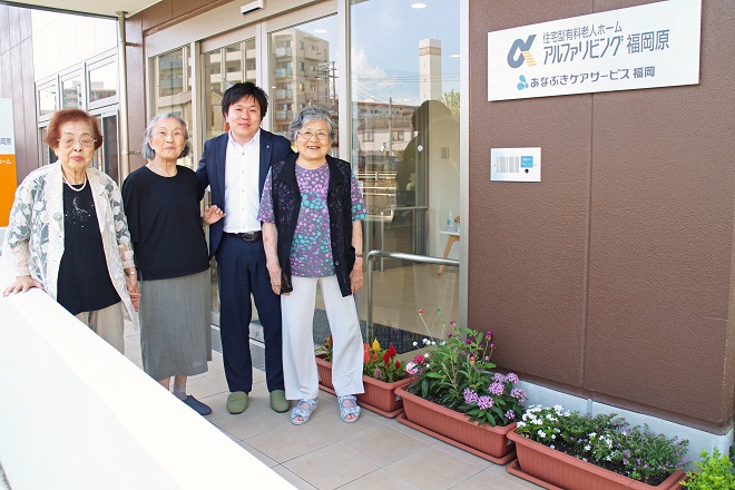 左側に入居者の３人と施設職員が見える，アルファリビング福岡原の正面玄関の写真