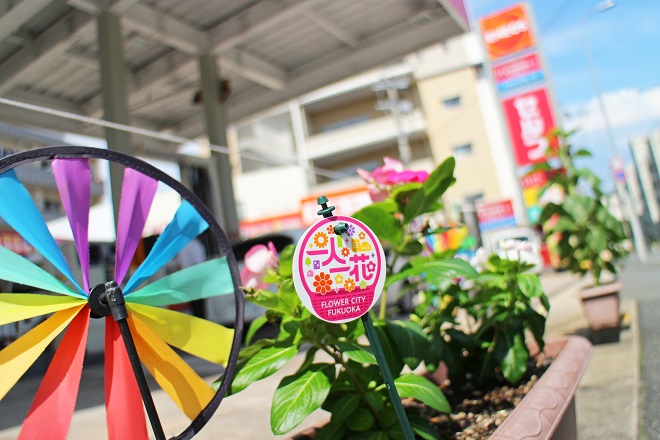 左に虹色の風車，中央に一人一花ロゴとピンクの花，右奥にスタンドの看板がぼけて見える写真