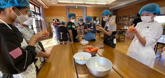 真剣に人参の皮をむくヘアキャップとエプロン姿の６年生の写真