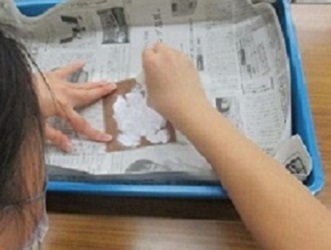 プラスティック引き出しに新聞紙を敷いた上に紙やすりを置い、石をこすりつけて削り出しているようす。