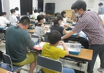 長机に二人ずつ座って一心に勾玉を削り出している参加者達の写真