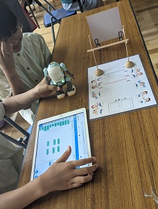 ロボットを左手に持ち右手でタブレットを操作している女子と、見ている姉妹の写真