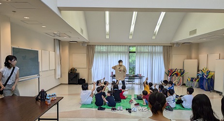 明るい窓を背に立つ講師の前に座って、話を聴いている20人ほどの子ども達