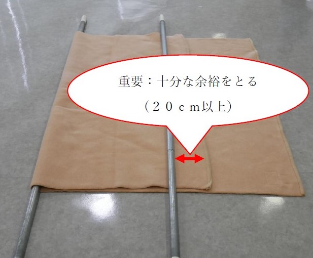 ４　毛布の上に２本目の棒を置く。２本目の棒を置く位置は，折り返した毛布の端から２０センチ以上内側。この時に，２本目の棒より外側で毛布の重なりが十分確保されているように注意する。