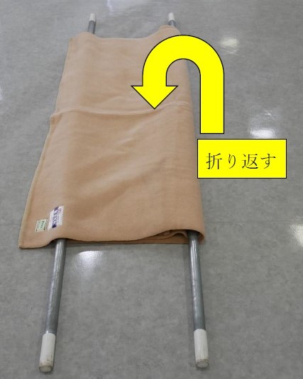 ５　２本目の棒を中心に，毛布の端末２枚をきれいに折り返す。折り返した毛布の端末は，１本目の棒を越えて，マチができるように注意する。