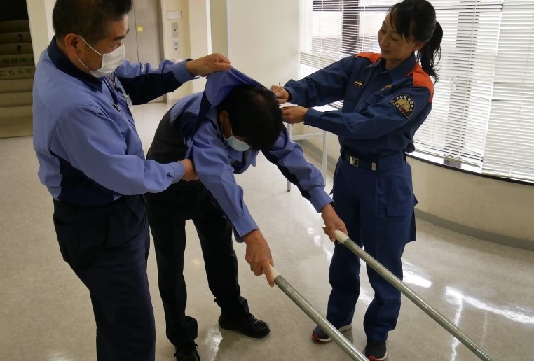 ２　搬送者１名が２本の棒の端末を左右の手でそれぞれつかむ。周囲の人が棒をつかんでいる人のシャツを下からめくりあげるように脱がしていく。この時，シャツのボタンは脱がせる際に頭が通る程度はずすが，その他のボタンはしっかりとめておく。脱がしたシャツは裏返しながら，つかまれている２本の棒に通していく。