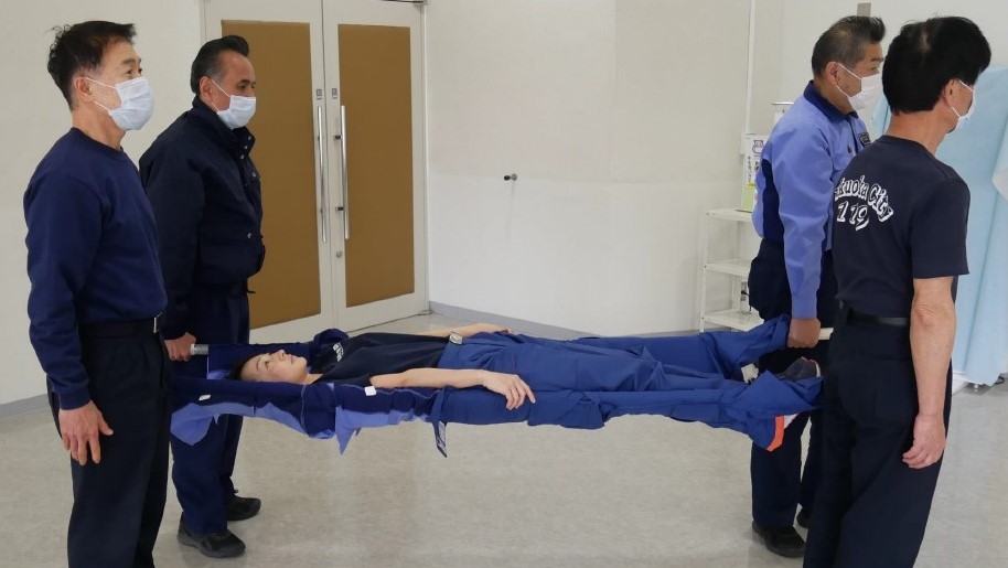 ４　傷病者を毛布の上に仰向けで寝かせ，２名から４名の搬送者がそれぞれ棒の端を持ち上げる。傷病者の足側を進行方向として搬送する。