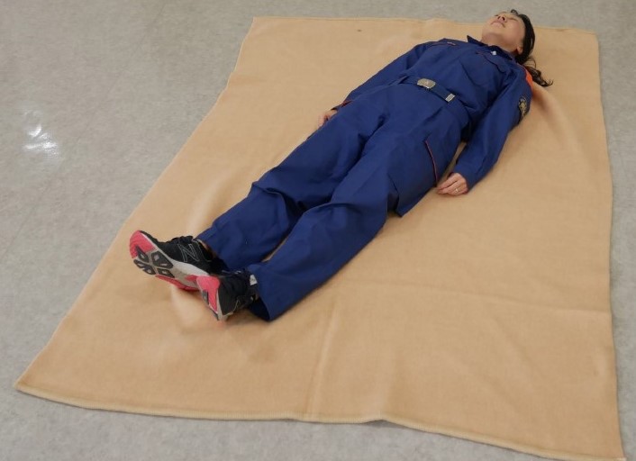 ２　傷病者を毛布の上に仰向けで寝かせる。寝かせる位置は，毛布の対角線上の中央で，傷病者の体が毛布からはみ出さないようにする。