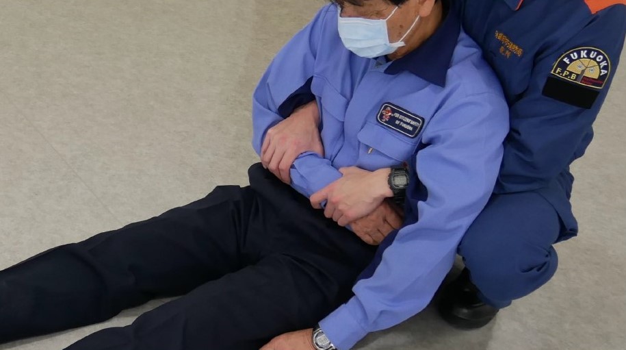 ２　傷病者の背中を支えたまま，両腕をそれぞれ両脇の下から通し，傷病者の片方の腕をつかむ。つかむ位置は，肘と手首の間をつかむ。