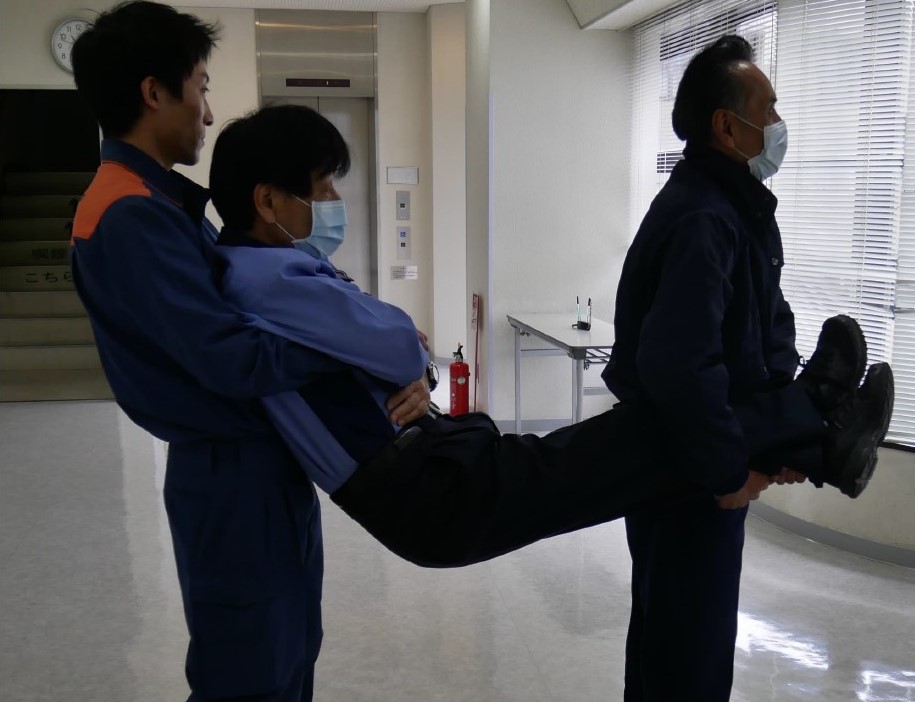 １　搬送者２名で傷病者を抱えている様子。１人目の搬送者は「背部から後方に移動する方法」と同じ要領で自分の胸の前で傷病者の腕を保持し，２人目の搬送者は交差させた傷病者の両足を，自分の腰の横で保持している。進行方向は傷病者の足側。