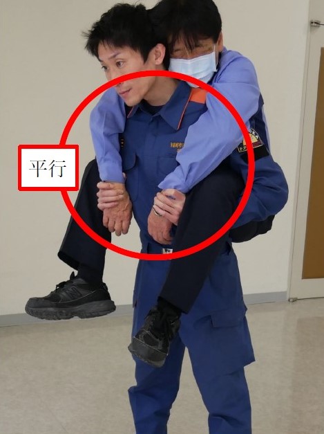 １　背負った傷病者の両腕を，搬送者の肩の上から，自然と下に垂らした状態で，搬送者が手首をつかんでいる様子。搬送者の腕は傷病者の太ももの下を通っている。