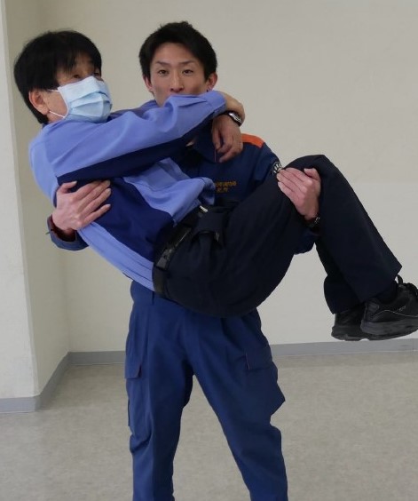 １　搬送者が，傷病者の背中を右腕で，両膝を左腕で抱え，お姫様抱っこの要領で抱えている様子。傷病者は両腕で，搬送者の首に両腕を回して捕まっている。