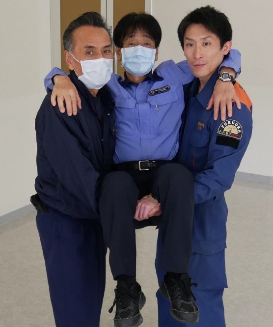 １　搬送者２名と傷病者１名が，横向きに並んでいる。並び順は搬送者，傷病者，搬送者の順番。３名はそれぞれ隣の人と肩を組んでいる。搬送者は，空いている方の手を傷病者の膝の下に通し，搬送者同士で手を繋ぐ。繋いだ手が腰の位置に来るように傷病者を抱え上げている。