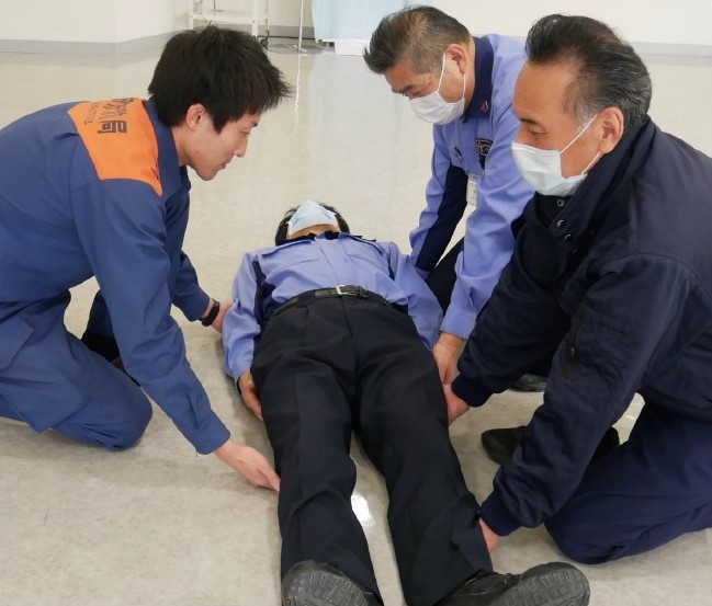 １　搬送者３名は仰向けになっている傷病者の両側に，交互に位置するように，片膝立ちの姿勢をとる。搬送者はそれぞれ，傷病者の背中の下に両腕を差し込む。